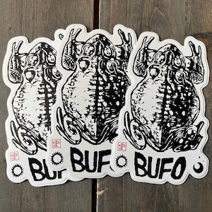 May include: Three white stickers with a black illustration of a toad. The stickers have the word "BUFO" in a bold, stylized font. The stickers are on a wooden surface.