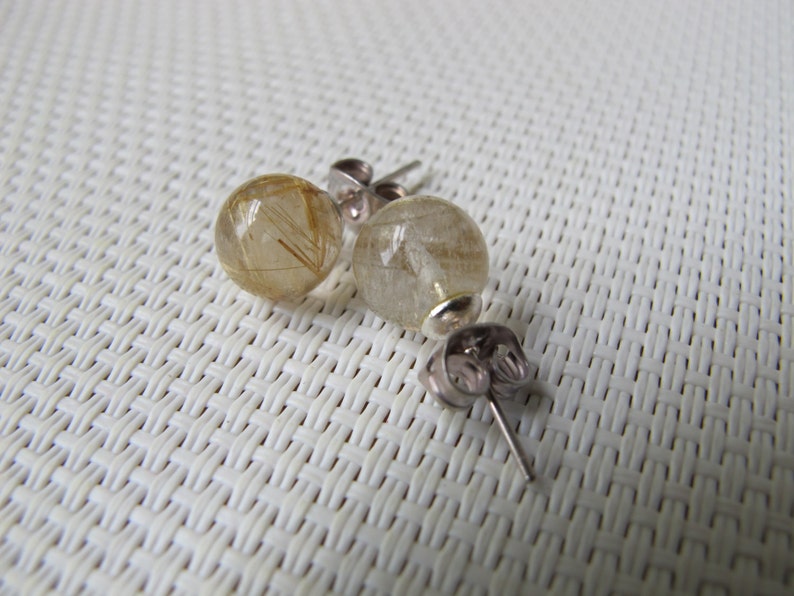 Pode incluir: Brincos de press&atilde;o com contas esf&eacute;ricas transparentes com inclus&otilde;es douradas em forma de cabelo. Os brincos t&ecirc;m pinos e fechos prateados. Apresentados em uma superf&iacute;cie branca texturizada.