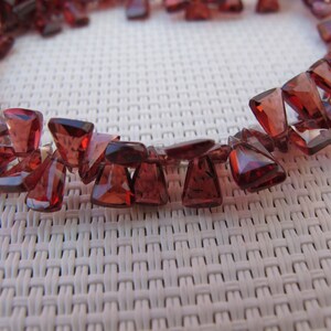 Puede incluir: Un collar compuesto por gemas triangulares facetadas de color rojo intenso. Las piedras están ensartadas, formando una forma curva. Las gemas tienen un aspecto pulido y translúcido, reflejando la luz. El collar se muestra sobre un fondo blanco tejido.