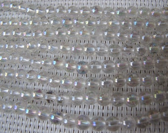 May include: Close-up of multiple strands of clear, iridescent beads. Each bead is round and appears to have a subtle rainbow sheen. The beads are strung together, creating a textured, shimmering effect. The background is a white woven surface.