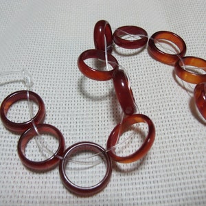 May include: A selection of translucent, reddish-brown rings threaded on a clear cord. The rings display a range of colours, from a deep burgundy to a lighter amber. The rings are likely made of polished stone or resin.