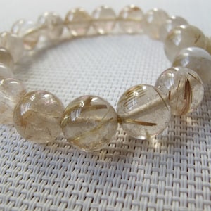 May include: A clear quartz bracelet with golden rutile inclusions. The round beads are strung together, displaying the natural patterns within the stones. The bracelet is on a white textured surface.