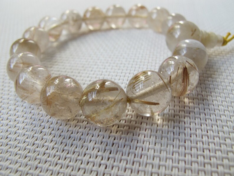 May include: A clear quartz bracelet with golden rutile inclusions. The round beads are strung together, displaying the natural patterns within the stones. The bracelet is on a white textured surface.