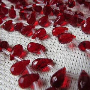 May include: Close-up of strands of faceted, teardrop-shaped red beads. The beads are strung on clear, thin cords, creating a delicate and elegant appearance. The beads are a deep, rich red color.