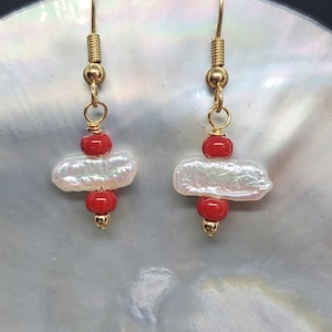 Puede incluir: Un par de pendientes de oro con cuentas rojas y blancas. Los pendientes presentan una cuenta blanca nacarada con una cuenta roja a cada lado.