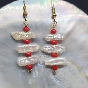 Può includere: Un paio di orecchini dorati con perle barocche bianche e perline di corallo rosso. Gli orecchini sono pendenti e hanno un design semplice.