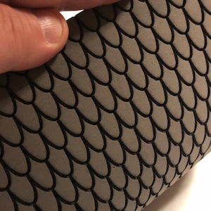 May include: Close-up of a gray, textured material with a scale-like pattern. The scales are outlined in black, creating a contrasting design. The material is being held by a hand, showcasing the texture and design.