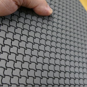 May include: Close-up of a gray, textured mat with a repeating, interwoven black pattern. The mat has a fishnet-like design, with a slightly raised texture. The mat is being held up by a hand, showing its flexibility and thickness.