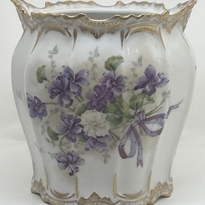 May include: A white porcelain cachepot featuring a hand-painted floral motif of purple violets and green leaves. The vessel has a scalloped rim and base, both accented with gold trim. A light purple ribbon is tied around the floral bouquet.