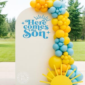 May include: A cream-colored sign with the text "Here comes the Son" in blue, decorated with a balloon arch in yellow, orange, and blue. A yellow sun cutout is at the bottom. The sign is likely for a party or event.