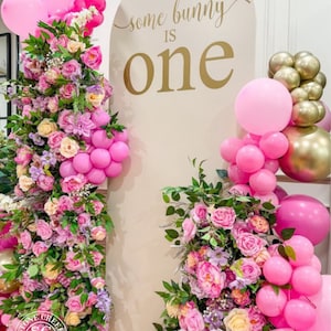 May include: A white backdrop with the text 'some bunny is one' in gold lettering. The backdrop is decorated with pink and gold balloons and pink and purple flowers.