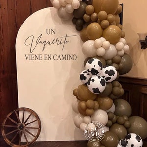May include: A white arched backdrop featuring the text UN Vaquerito VIENE EN CAMINO in black lettering. A balloon garland in shades of brown, cream, and white with cow-print patterns is attached to the side. A wooden wagon wheel sits on the floor.