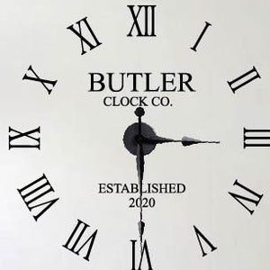 May include: A black and white clock decal with Roman numerals and the text "BUTLER CLOCK CO. ESTABLISHED 2020". The decal is applied to a white wall above a gray couch with black and white patterned pillows.