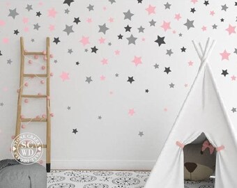 Puede incluir: Una tienda de campaña tipi blanca con lazos rosas en la entrada, instalada en una habitación con una pared blanca decorada con pegatinas de estrellas rosas y grises. Un puf gris se encuentra delante de la tienda de campaña.