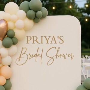 May include: A white arched backdrop sign with the gold text PRIYA'S Bridal Shower, decorated with a balloon garland in shades of cream, sage green, peach, and metallic gold, including three heart-shaped balloons and a small lantern nearby.
