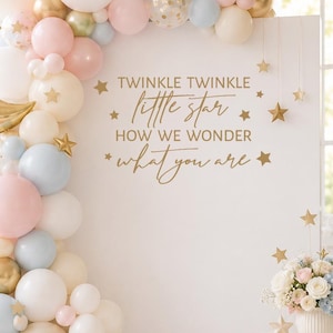 May include: A white backdrop featuring the gold text TWINKLE TWINKLE little star HOW WE WONDER what you are, surrounded by a balloon arch in white, pink, blue, and gold, a gold crescent moon balloon, a teddy bear, and a floral arrangement.