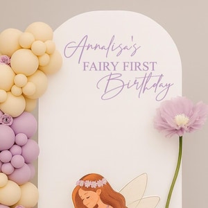 May include: A white arched backdrop featuring the purple text Annalisa's FAIRY FIRST Birthday, decorated with a balloon garland in shades of cream, lavender, and light pink, a fairy illustration, and a large artificial purple flower.