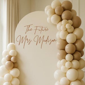 May include: A large arched backdrop in a solid cream color featuring the text The Future Mrs. Madison in a brown script font. The backdrop is decorated with a balloon garland in shades of cream, beige, and tan, arranged along the side and top.