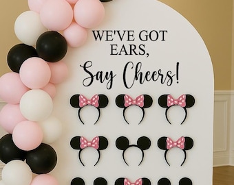 May include: A white arched display board features the black text WE'VE GOT EARS, Say Cheers! and twelve black mouse ear headbands, some with pink polka-dot bows. A balloon garland in pink, white, and black wraps around the left side of the board.