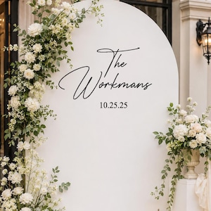 May include: A large white arched backdrop featuring the black text The Workmans and the date 10.25.25. The display is decorated with white floral arrangements, green foliage, and several gold-framed lanterns containing white pillar candles.