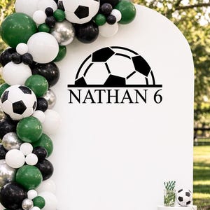 May include: A soccer-themed party decoration featuring a white arch with the name "NATHAN 6" and a soccer ball graphic. The arch is decorated with a balloon garland in green, black, white, and silver. A large soccer ball is on the ground.