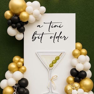 May include: A white rectangular backdrop featuring the black text 'a Tini bit older' and a large martini glass illustration with three green olives. The display is decorated with clusters of gold, white, and black balloons on both sides.