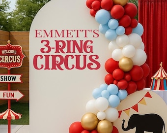 May include: A large white arched backdrop with the red text EMMETT'S 3-RING CIRCUS, decorated with a balloon garland in red, white, light blue, and gold. Includes a red circus sign, a small circus tent, a black elephant silhouette, and a red drum.