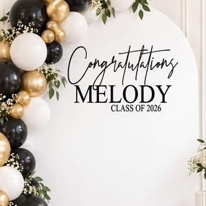 May include: A white arched backdrop featuring black text that reads Congratulations MELODY CLASS OF 2026. A balloon garland in black, white, and gold with greenery surrounds the arch. A graduation cap sits on a stack of books with a diploma.