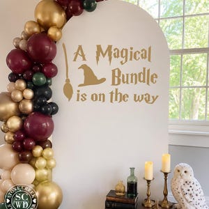 May include: A white arched backdrop featuring the gold text A Magical Bundle is on the way with a broom and hat graphic. A balloon garland in burgundy, gold, dark green, and beige accents the side. Includes a stack of books, bottles, candles, and an owl.