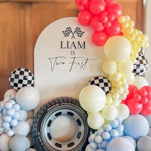 May include: A white arched backdrop featuring the text LIAM IS Two Fast with two checkered flags, surrounded by a balloon garland in red, yellow, light blue, and white, with checkered balloons and a black rubber tire with a grey rim.