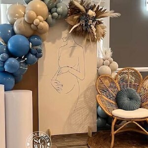 May include: A tall rectangular backdrop featuring a black line art drawing of a pregnant person, decorated with a cluster of blue and beige balloons and a dried floral arrangement. A rattan peacock chair with a blue round cushion sits nearby.