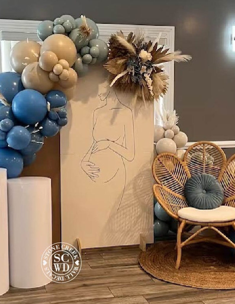 May include: A tall rectangular backdrop featuring a black line art drawing of a pregnant person, decorated with a cluster of blue and beige balloons and a dried floral arrangement. A rattan peacock chair with a blue round cushion sits nearby.
