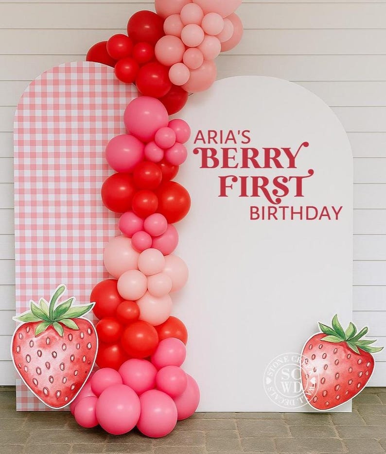 May include: A festive birthday backdrop featuring a white arch with the text "ARIA'S BERRY FIRST BIRTHDAY" in red. A balloon garland in shades of red and pink adorns the arch. Two large strawberry cutouts flank the backdrop, and a pink and white gingham panel is on the left.