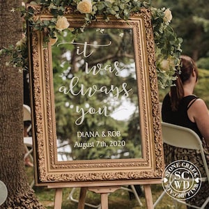 May include: A gold-framed mirror with a white vinyl decal that reads "it was always you" with the names Diana & Rob and the date August 7th, 2020. The mirror is standing on an easel in front of a green leafy background.