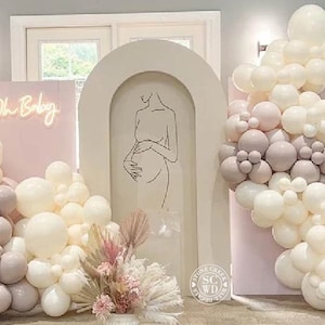 May include: A beige arched backdrop featuring a black line art illustration of a pregnant person. The display includes a pink panel with a neon Oh Baby sign, clusters of white and mauve balloons, and a floral arrangement of dried pampas grass.