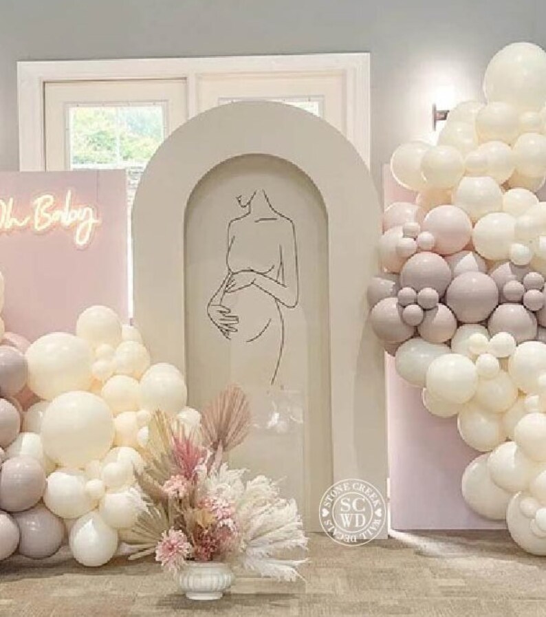 May include: A beige arched backdrop featuring a black line art illustration of a pregnant person. The display includes a pink panel with a neon Oh Baby sign, clusters of white and mauve balloons, and a floral arrangement of dried pampas grass.