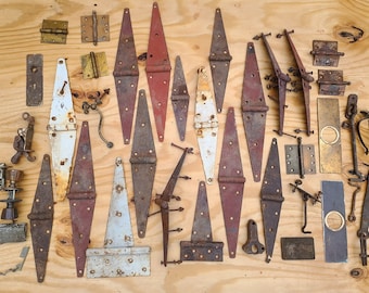 May include: A collection of vintage metal hinges, latches, and hardware in various sizes and styles. The pieces display a range of colors, including red, white, and shades of brown, with visible signs of age and wear. The hardware is arranged on a light wood surface.