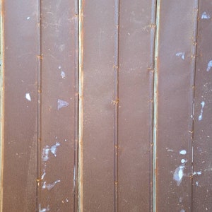 May include: A close-up of a rusty metal wall with vertical panels. The wall is a dark brown color with some white paint splatters.