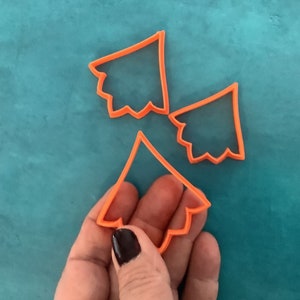May include: Three orange plastic cookie cutters shaped like Christmas trees. The cutters are on a teal surface.