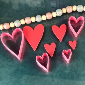 May include: A set of eight pink heart-shaped cookie cutters of varying sizes. The cutters are arranged on a dark green surface.