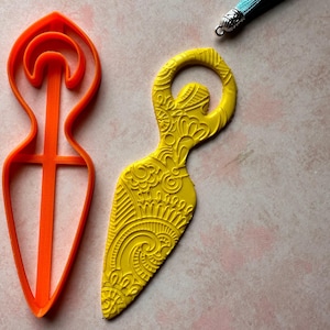 May include: An orange cookie cutter and a yellow decorative item with an intricate floral design are displayed on a pink surface. A blue tassel with a silver cap is also present. The items appear to be craft or baking related.