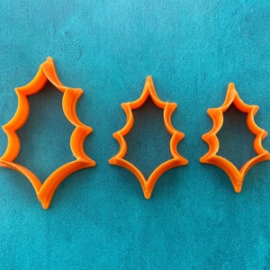 May include: Three orange plastic holly leaf-shaped cookie cutters of varying sizes. The cutters have a jagged edge and are arranged from largest to smallest. The background is a solid teal color.