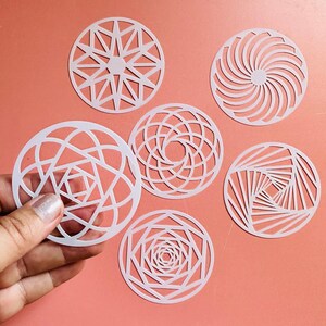 May include: A collection of six white, circular decorative pieces with intricate geometric designs. The designs include starbursts, spirals, and repeating patterns. The pieces are displayed on a peach-colored surface.