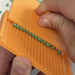 Puede incluir: Una herramienta de plástico naranja con una serie de ranuras para alinear pequeñas cuentas verdes. La herramienta está siendo sostenida por las manos de una persona.