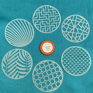 May include: Six white plastic stencils with different geometric designs. The stencils are on a turquoise background. The stencils are for crafting and decorating.
