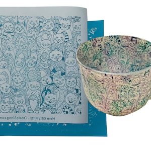 Puede incluir: Una plantilla azul con el texto "Here kitty kitty - CreateAlong.com" y un diseño de muchos gatos. Se muestra una taza de cerámica blanca con un diseño de gato similar en el fondo.