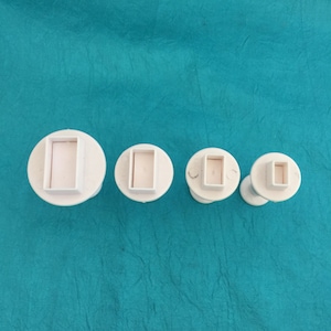 Puede incluir: Cuatro cortadores de galletas rectangulares de pl&aacute;stico blanco de diferentes tama&ntilde;os. Los cortadores est&aacute;n sobre una superficie azul.
