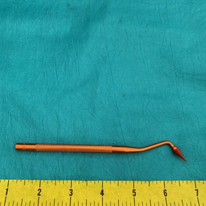 May include: A close-up of a small, orange-colored dental tool with a pointed tip. The tool is about 6 inches long and has a textured handle. A yellow ruler is visible in the background.