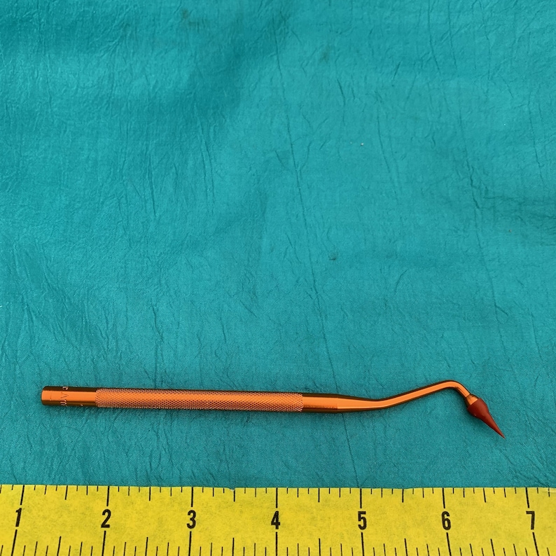 May include: A close-up of a small, orange-colored dental tool with a pointed tip. The tool is about 6 inches long and has a textured handle. A yellow ruler is visible in the background.