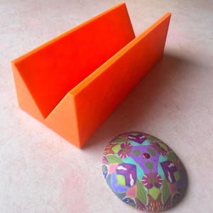 Può includere: Un supporto per telefono in plastica arancione con una base triangolare e uno slot per tenere un telefono. Un oggetto rotondo e colorato con motivi è in primo piano.